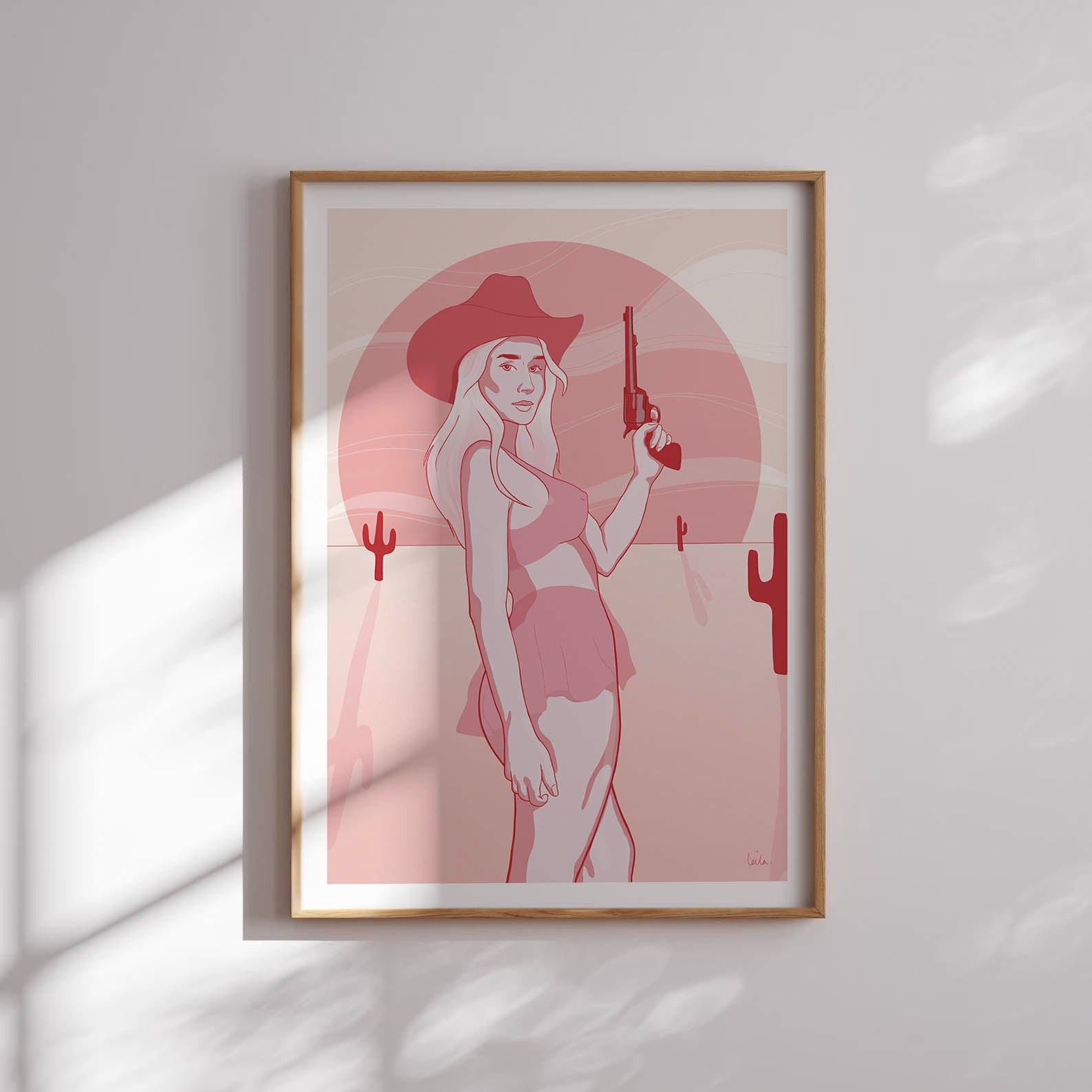 A framed poster depicting a digital drawing of a pink cowgirl in the desert, placed against a white wall.