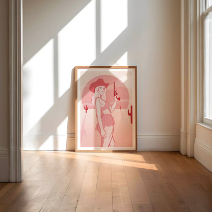 A framed poster depicting a digital drawing of a pink cowgirl in the desert, placed against a white wall.